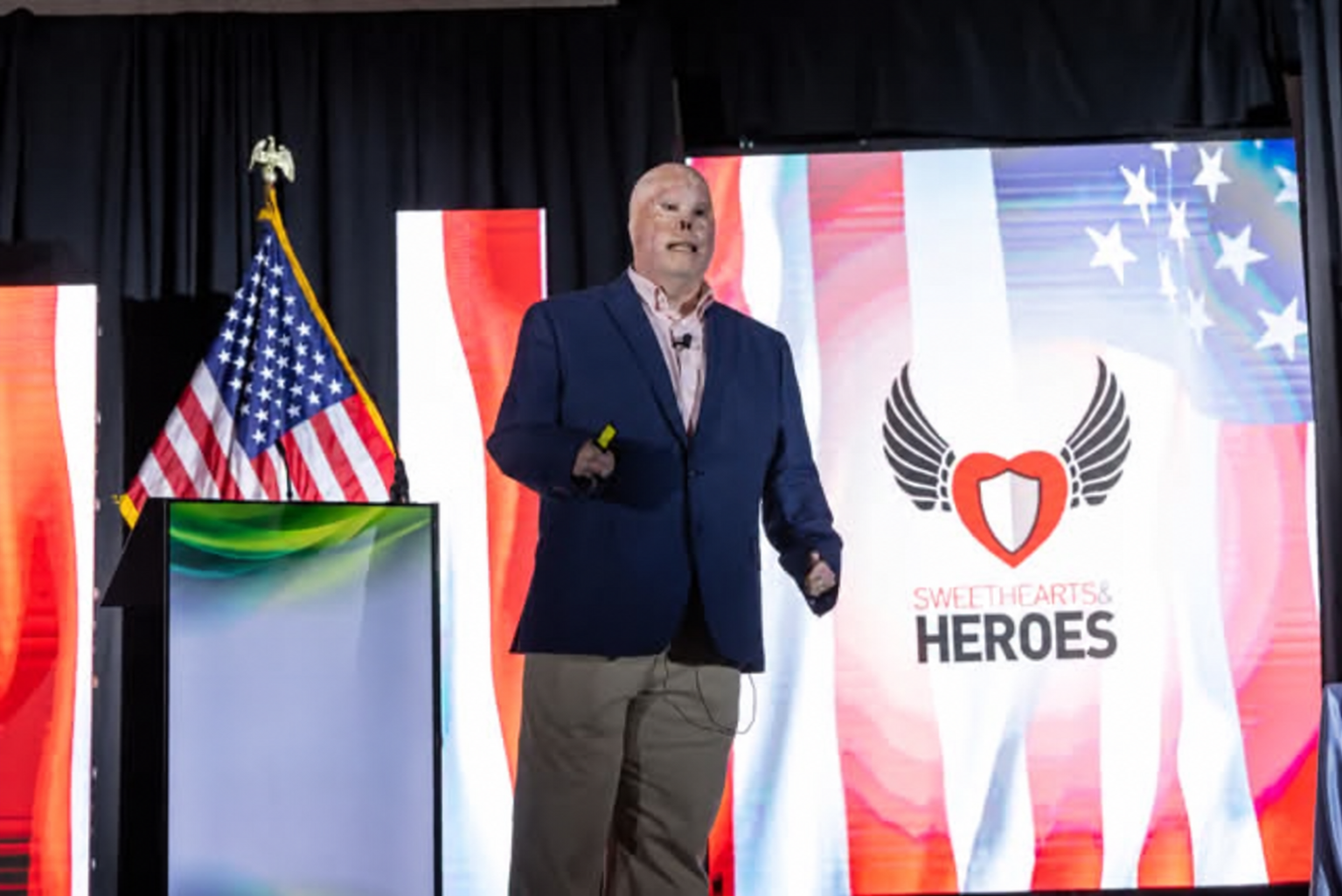 Rick Yarosh speaking on stage in a blue blazer at a Sweethearts & Heroes event